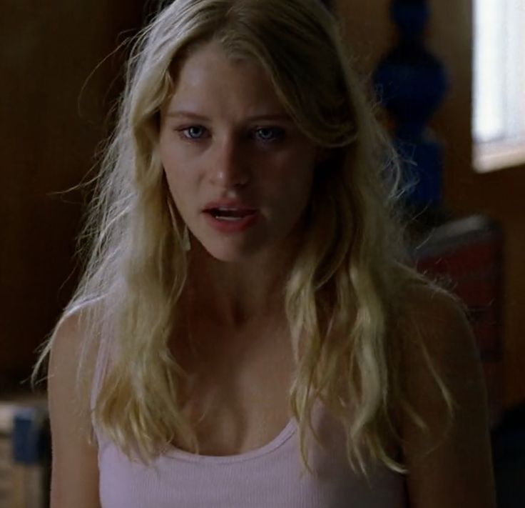 a woman with blonde hair and blue eyes in a white tank top looking at the camera