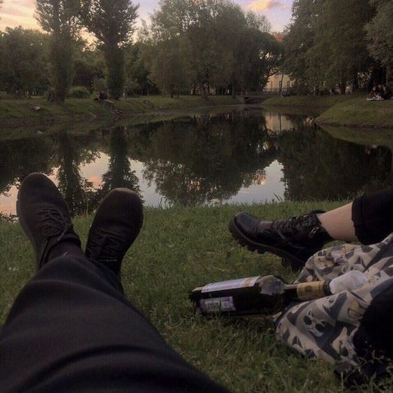 two people sitting next to each other on the grass near a pond with trees in the background