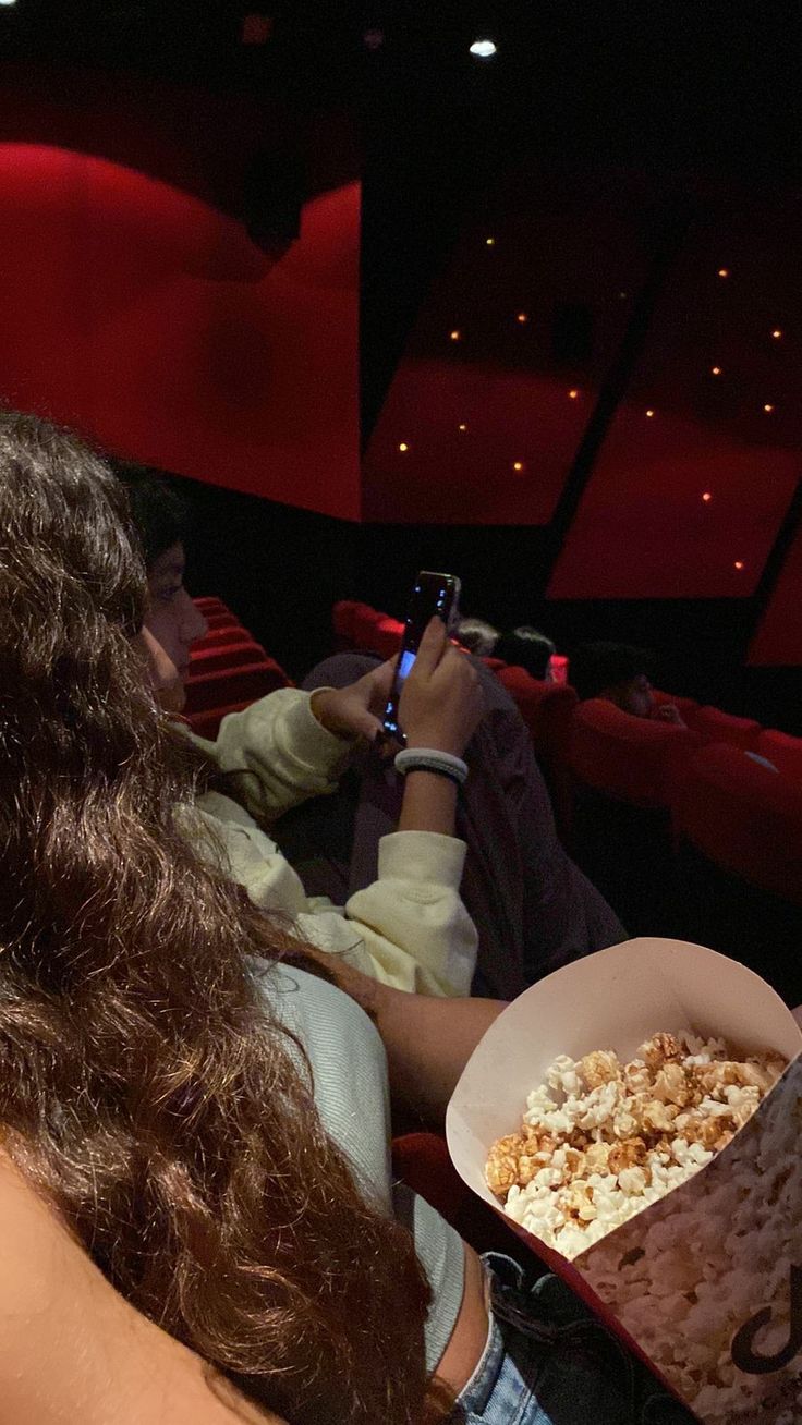 two people sitting in a movie theater and one is holding a box of popcorn while the other holds a cell phone