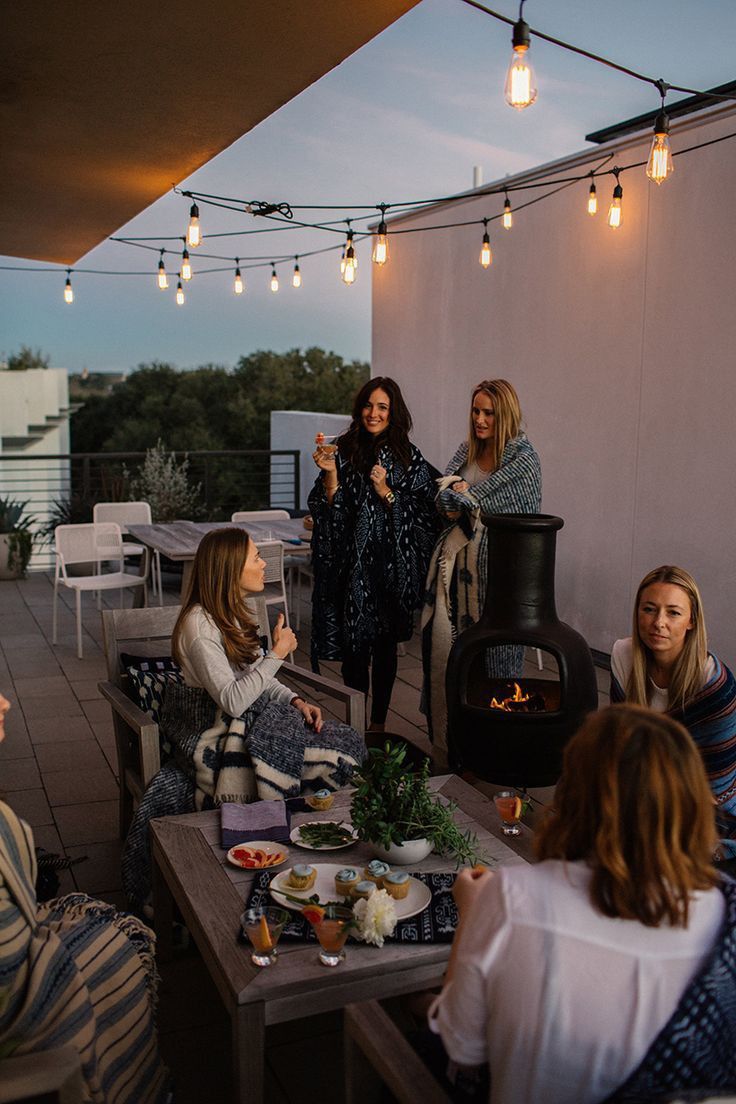 several women sitting around a table with food and drinks in front of an outdoor fireplace