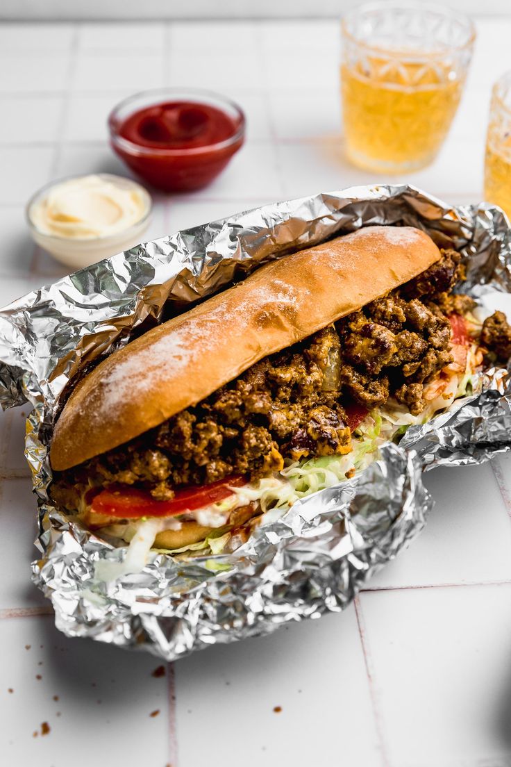 a meat sandwich sitting on top of tin foil next to some cups and sauces