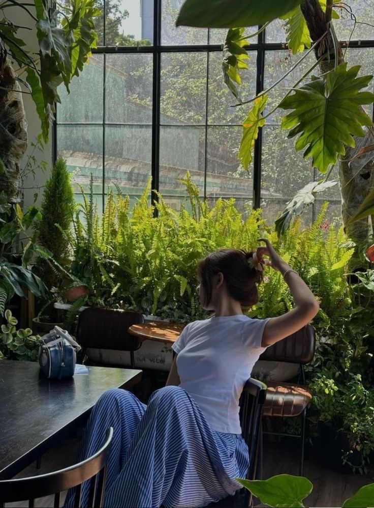 a woman is sitting at a table in front of plants and looking out the window