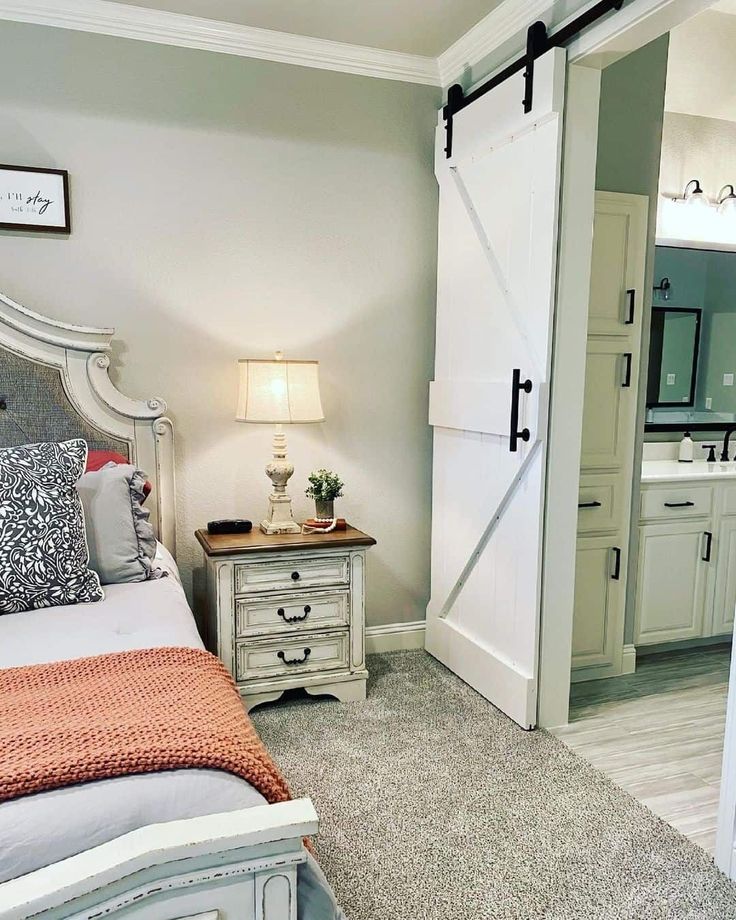 An elegant white and gray French bed fitted with gray and orange bedding is set in front of an ensuite bath. The bathroom is finished with a white sliding barn door on black metal tracks. Add rustic charm with a distressed two-toned white night table lit by a white and beige table lamp...   Image: 4y_farmhouse Lights At Night, Pantry Door, The Christmas, At Night, Pantry, Night Light, Christmas Decor, Instagram Profile, Trees