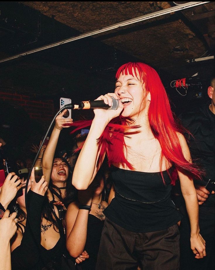 a woman with long red hair singing into a microphone at a music concert while people look on