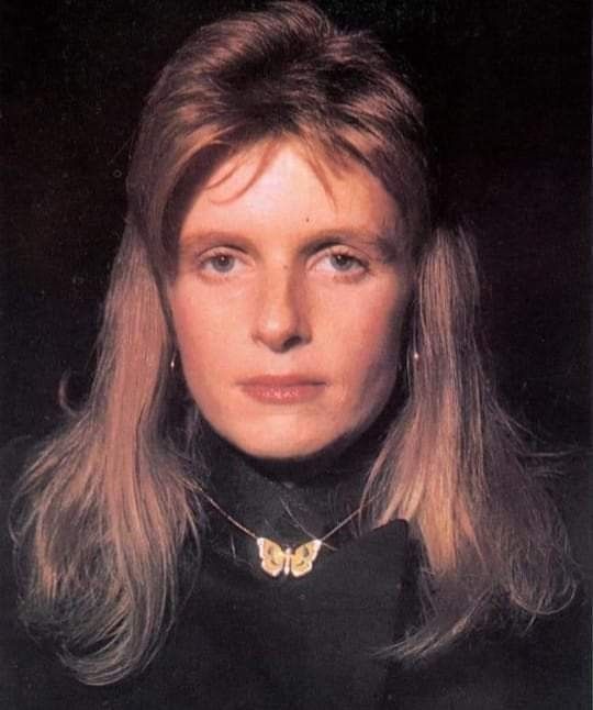 a woman with long blonde hair wearing a black shirt and gold butterfly choker necklace