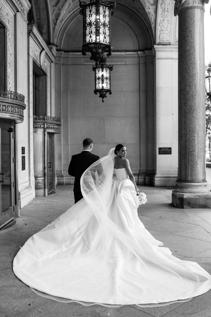 the bride and groom are walking through an old building with columns, chandeliers and windows