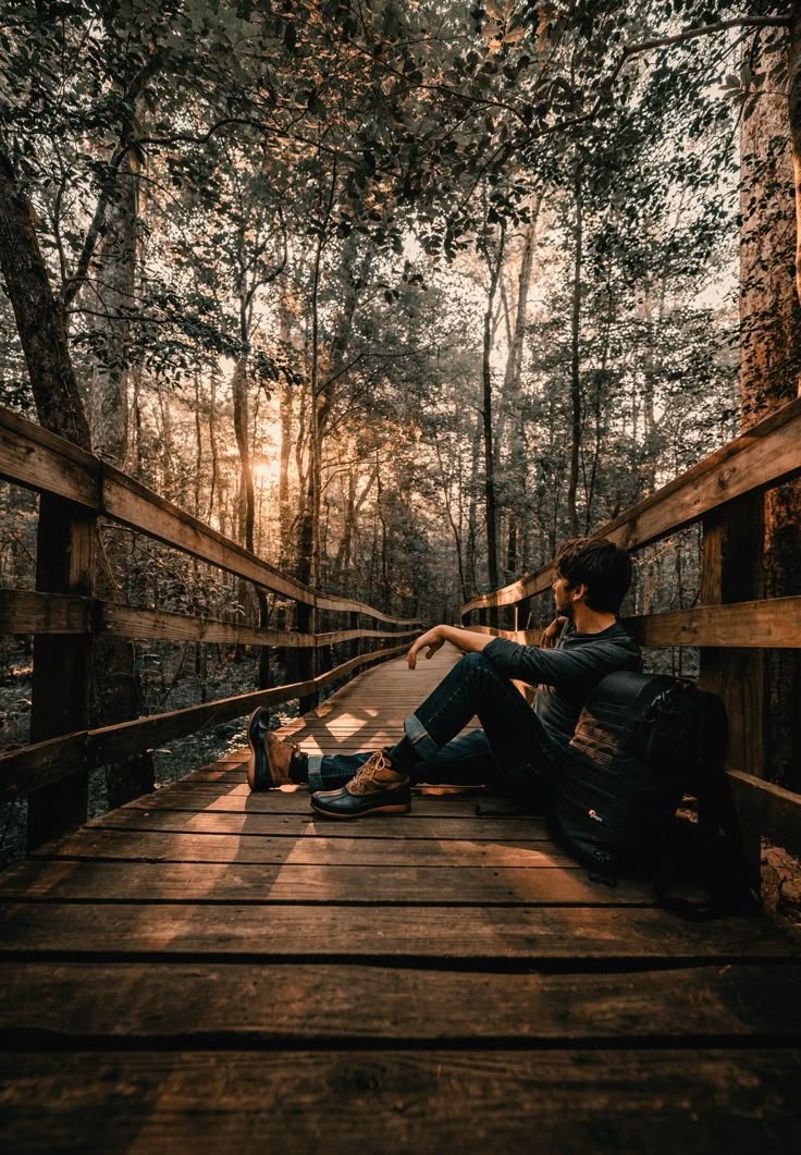 a man sitting on a wooden bridge in the woods with his feet crossed and looking at something