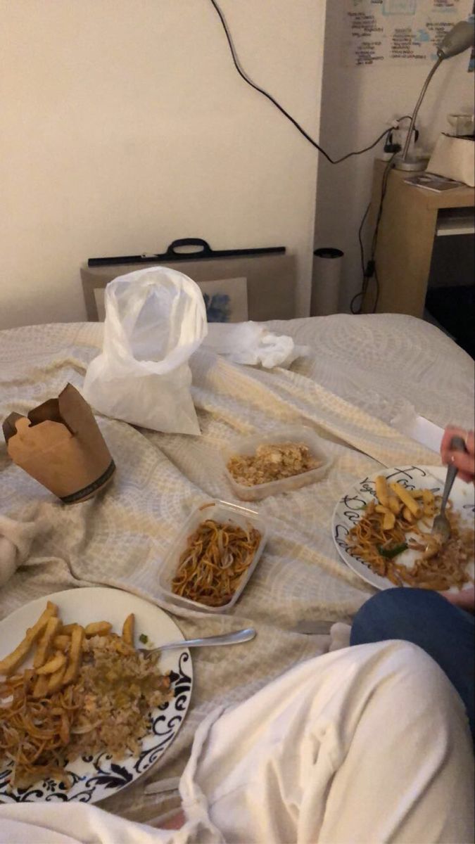 a person sitting on a bed with plates of food in front of them and a bag of chips to the side