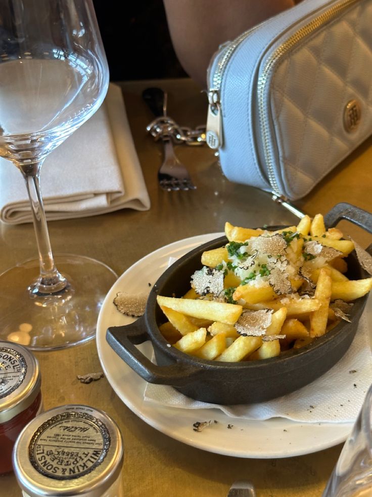 french fries in a skillet with cheese and herbs on the side next to some wine glasses