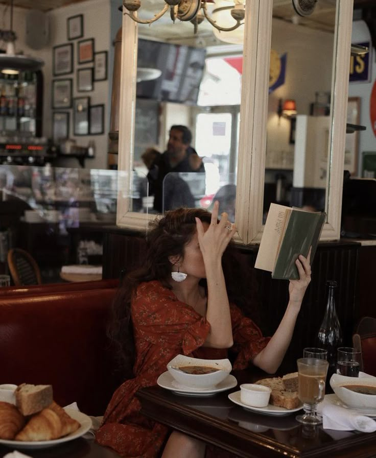 a woman reading a book while sitting at a table in a restaurant with food on it