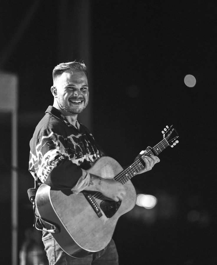 a man holding a guitar in his right hand and smiling at the camera while standing on stage