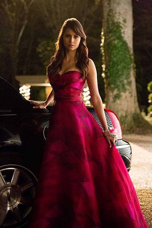 a woman in a long red dress standing next to a car and looking at the camera