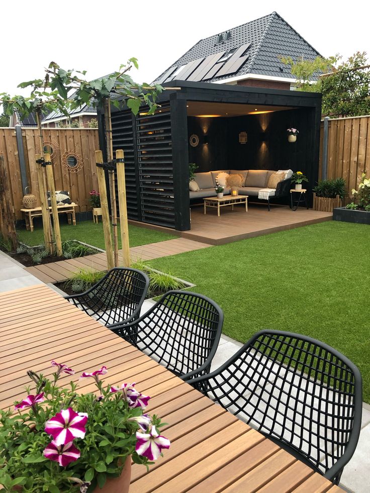 an outdoor living area with wooden tables, chairs and flowers on the grass in front of it