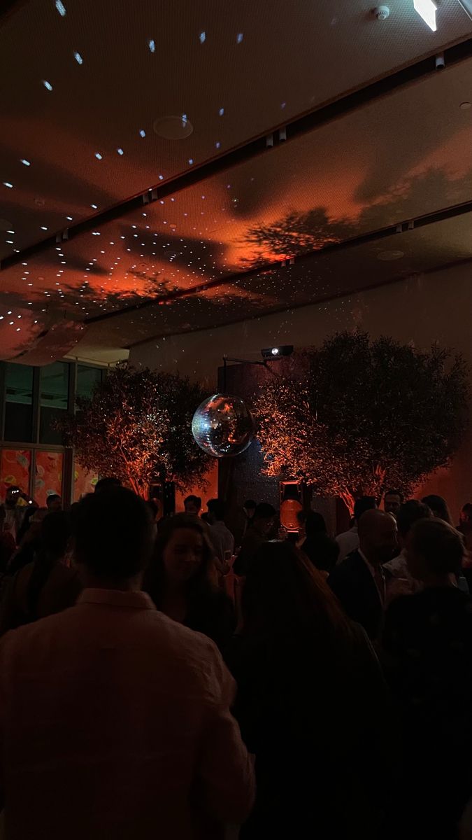 a large group of people in a room with lights on the ceiling and some trees