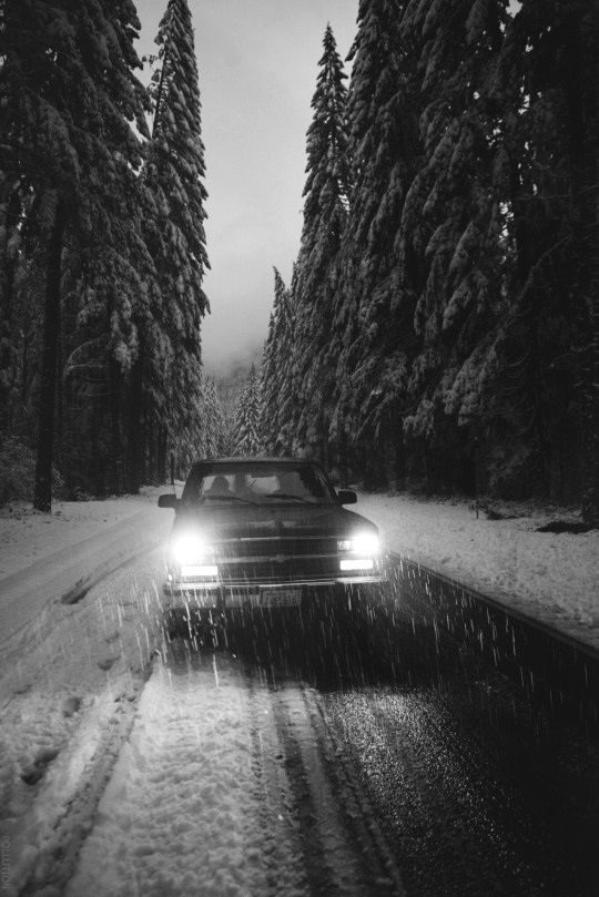 a car driving down a snow covered road at night with the headlights on and trees in the background