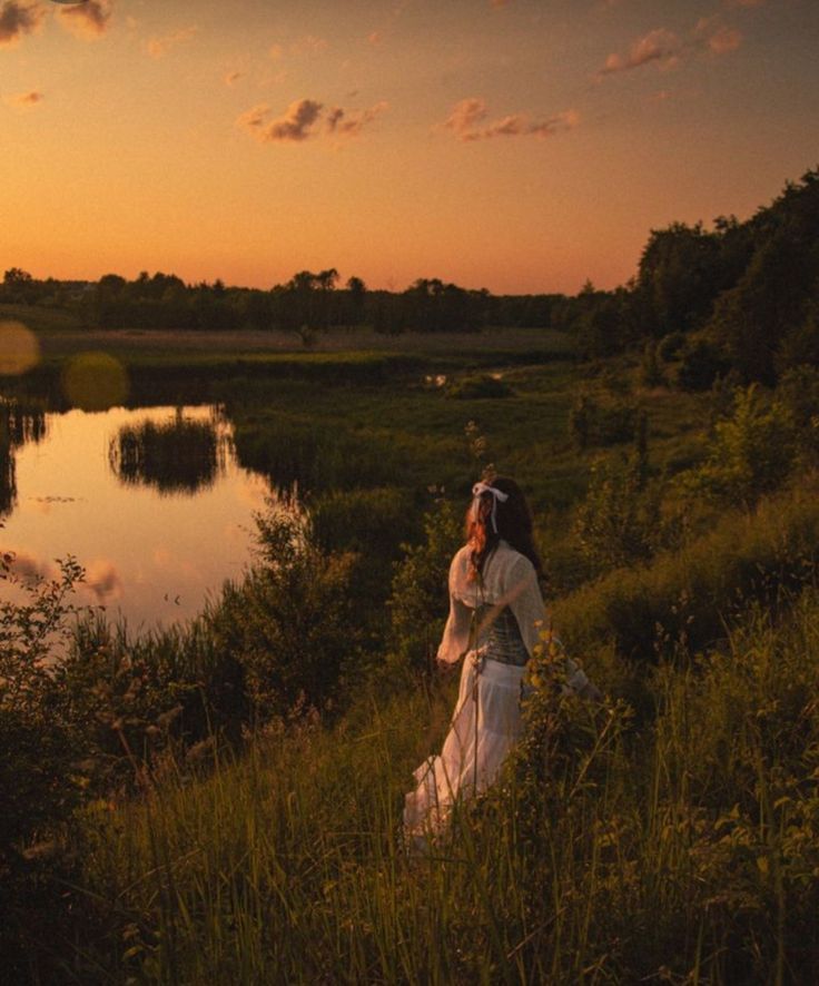 a woman in a white dress is standing by the water at sunset with her back to the camera