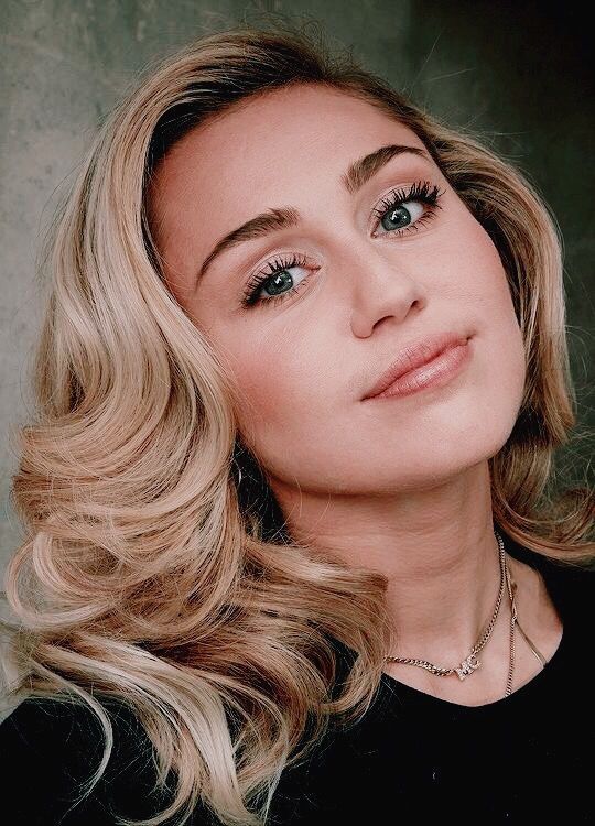 a woman with long blonde hair wearing a black shirt and necklace looking at the camera