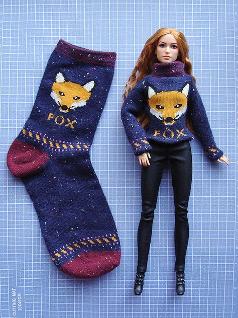 a doll is posed next to a knitted sock with a fox on the front