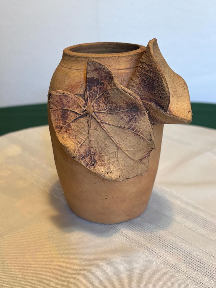 a brown vase with a leaf on it sitting on a white cloth covered tablecloth