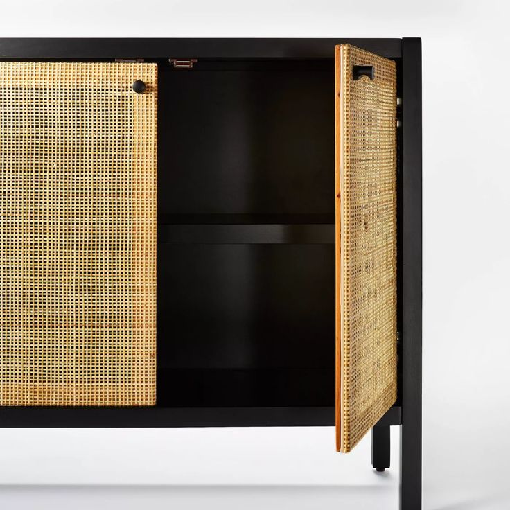 an open cabinet with wicker doors and black frame on the bottom, in front of a white wall