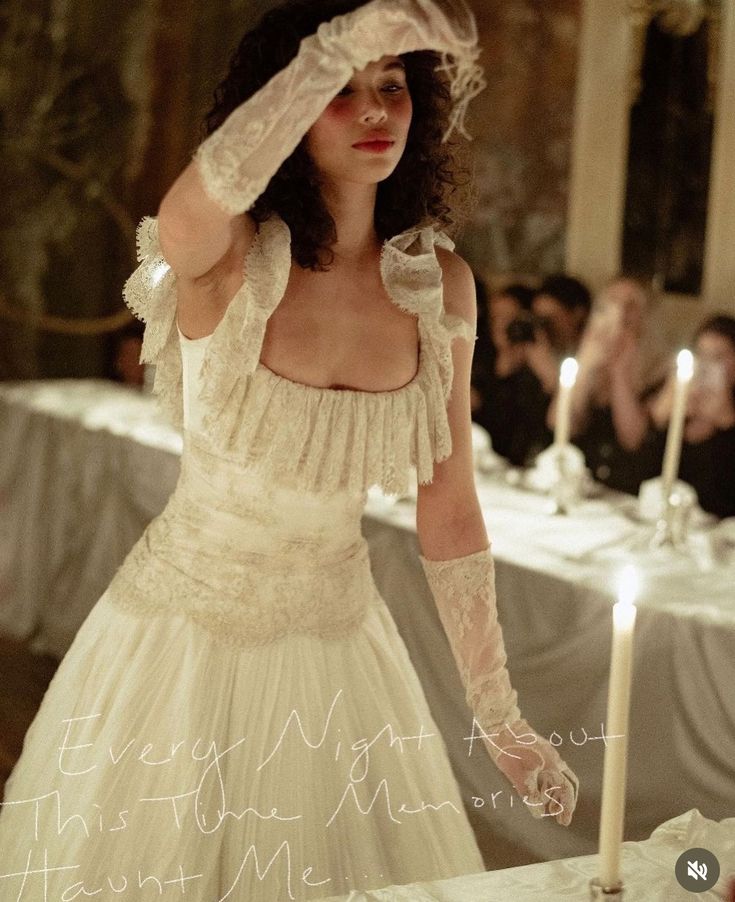 a woman in a white dress standing next to a table with two candles on it