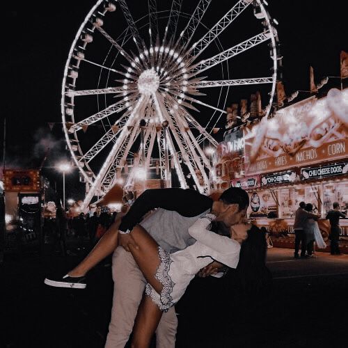 a man and woman kissing in front of a ferris wheel at night with lights on