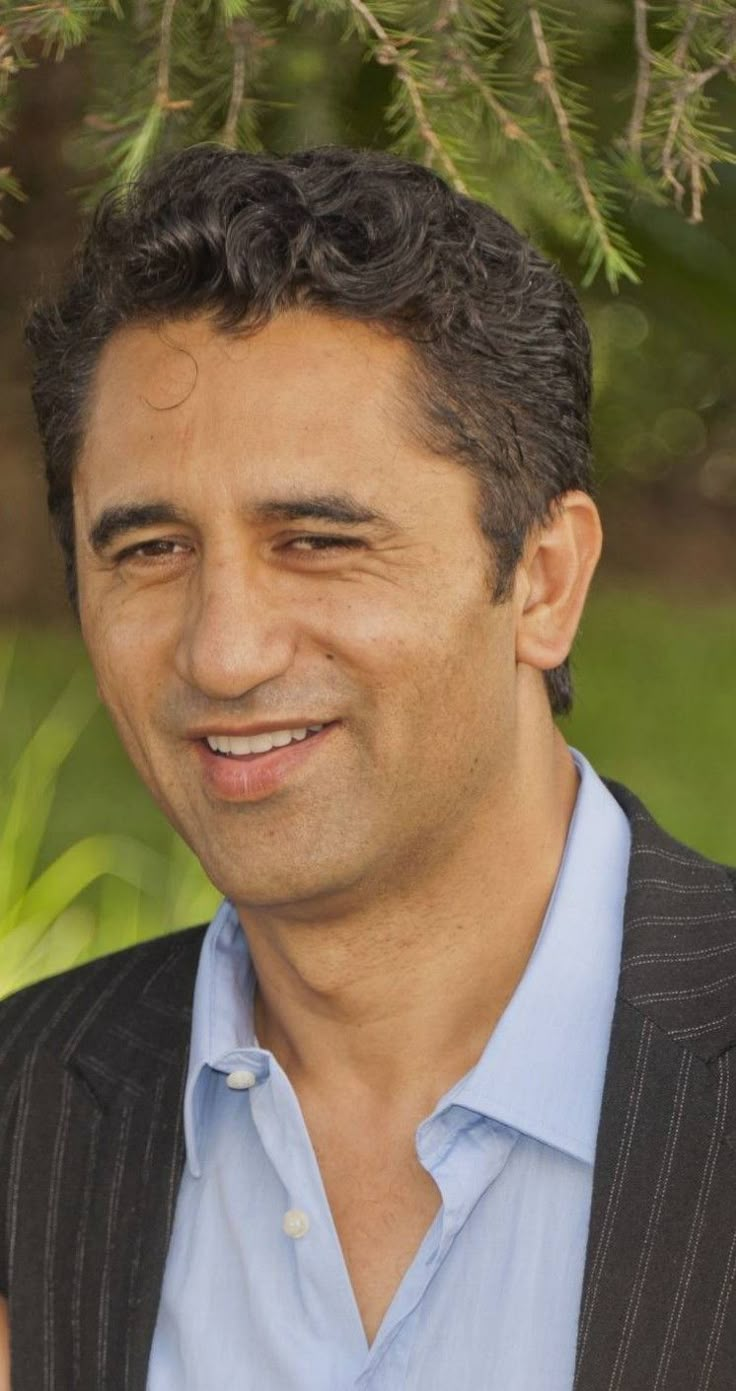 a man in a suit and blue shirt smiling at the camera with trees in the background