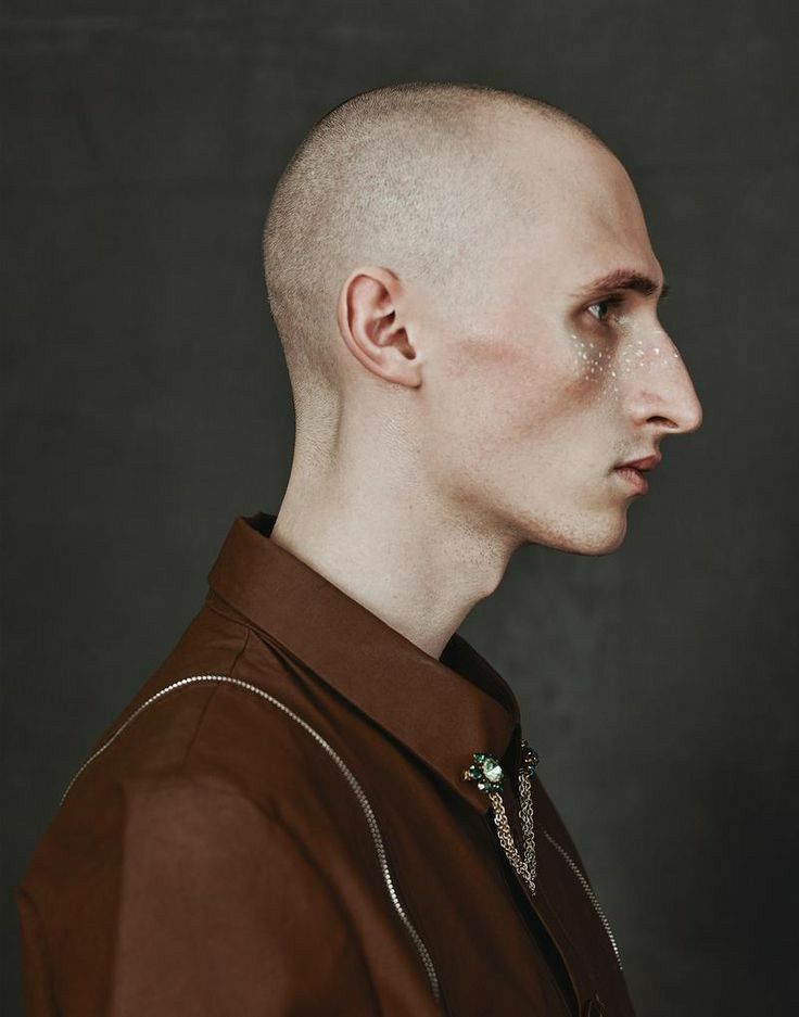 a bald man in a brown shirt and necklace looking off to the side with his head slightly shaved