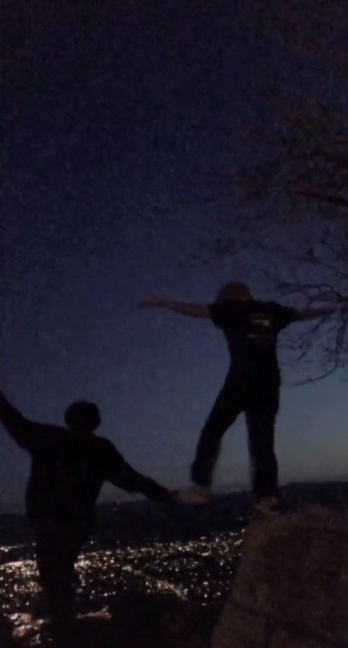 two people standing on the edge of a cliff at night with their arms in the air