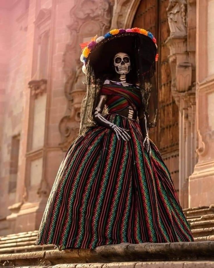 a skeleton in a mexican dress and sombrero stands on the steps outside an old building