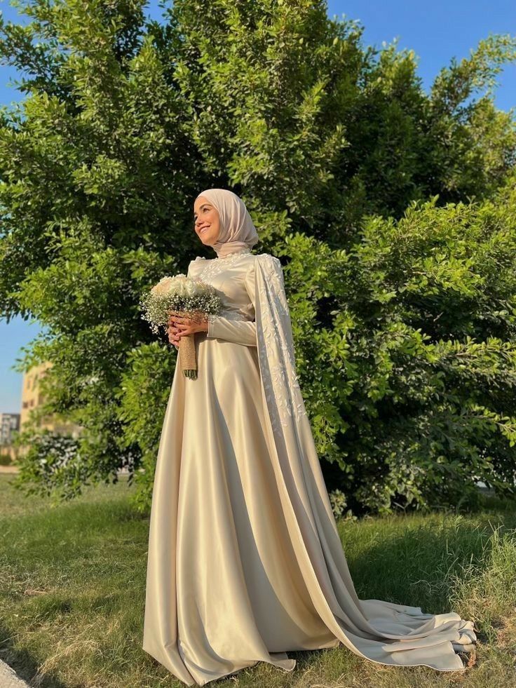 a woman in a white dress and veil holding a bouquet of flowers standing next to a tree