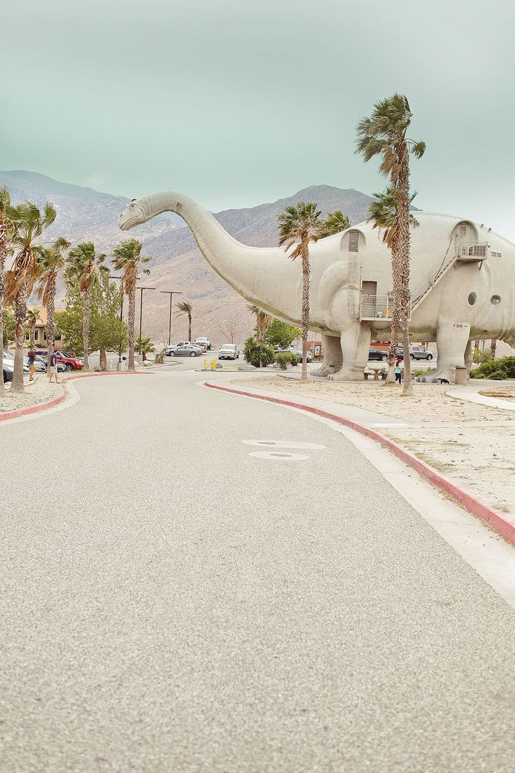 a large dinosaur statue sitting on the side of a road next to palm trees and mountains