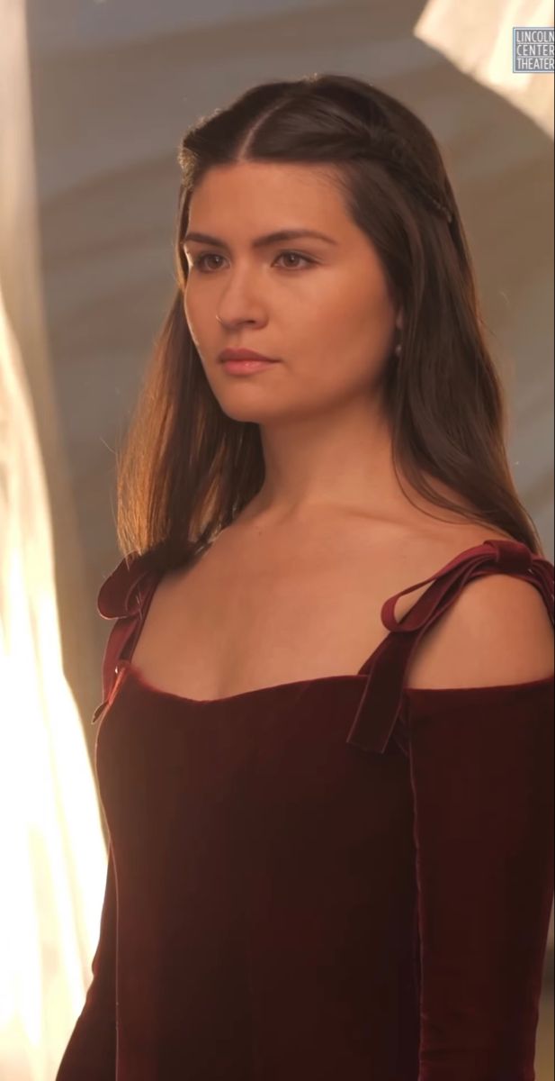 a woman with long hair wearing a red dress and looking off to the side while standing in front of a curtain