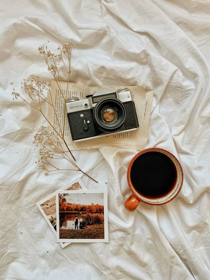 an old camera, coffee cup and photos are laying on a white sheet with some dried flowers