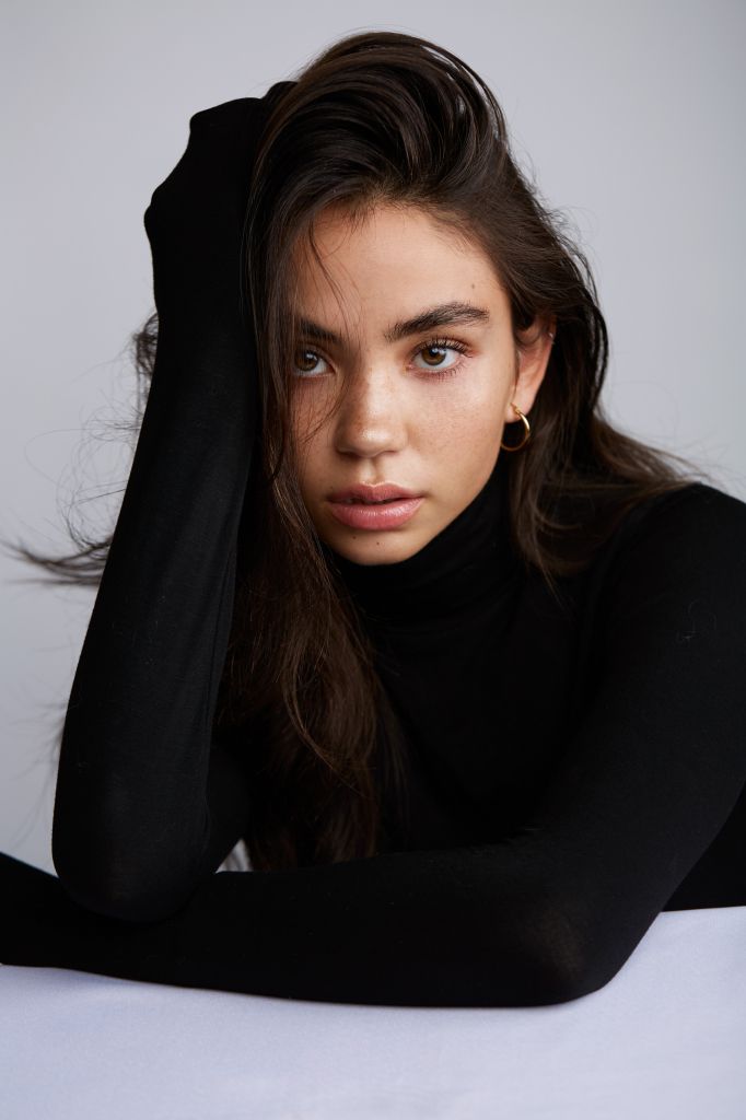 a woman sitting on top of a bed with her hands behind her head and looking at the camera