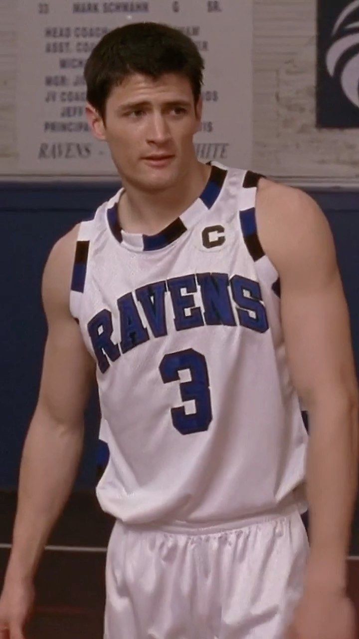 a young man standing in front of a basketball court wearing a white and blue uniform