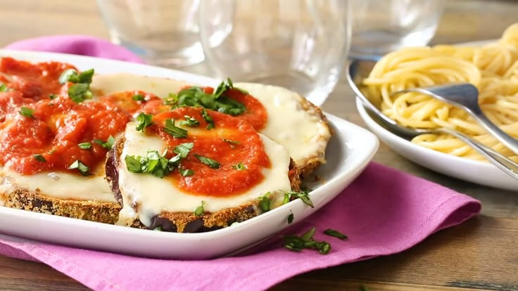 a white plate topped with meat covered in sauce and cheese next to pasta on a pink napkin