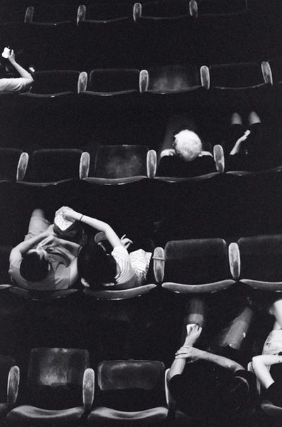 black and white photograph of woman laying on the seats in an empty theater auditorium with people watching