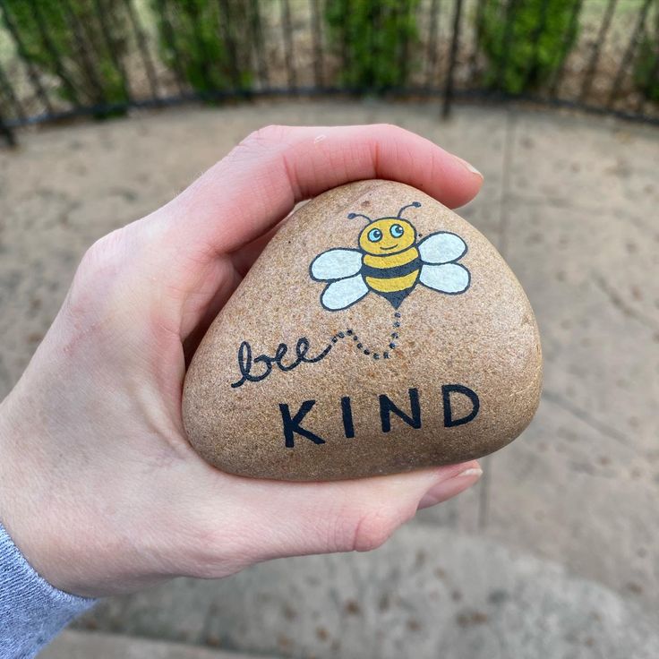 a hand holding a rock that says be kind with a bee painted on the side