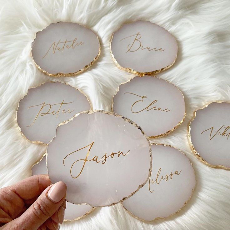 six personalized glass coasters with gold lettering on white furnishing, one being held up by a person's hand