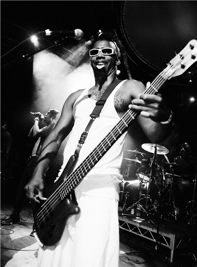 a man with sunglasses on holding a bass in front of him and another musician behind him