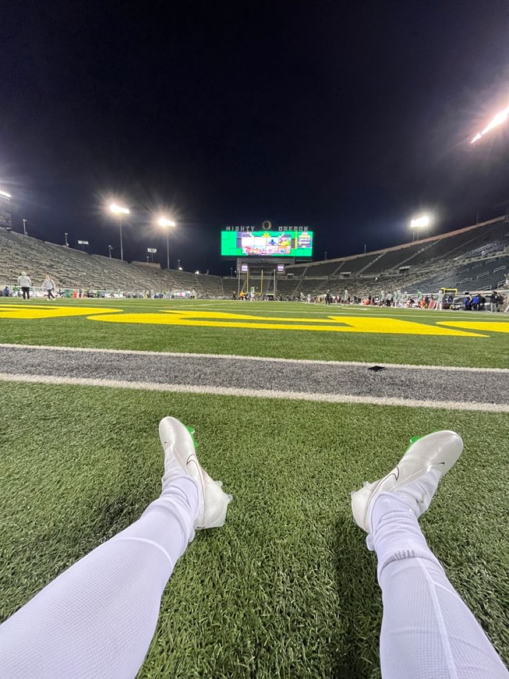 a person's feet resting on the grass at a football stadium with lights in the background