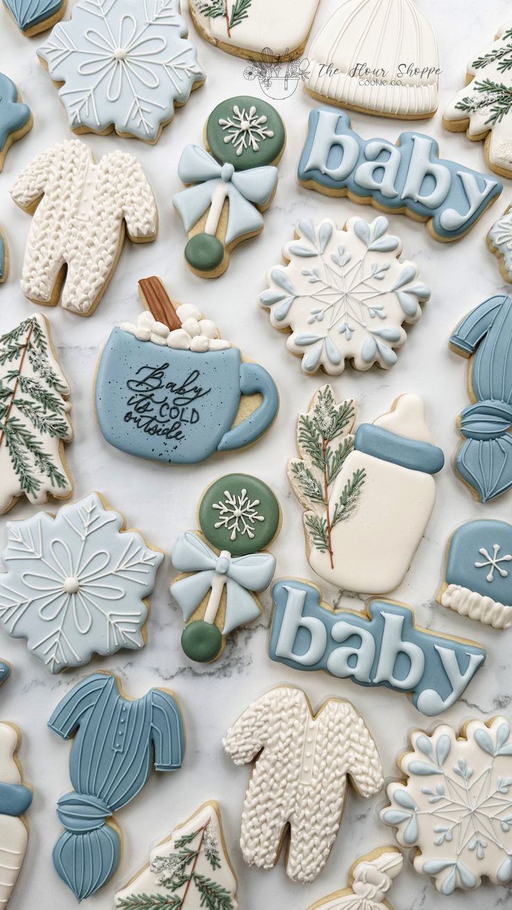 baby shower cookies are arranged on a white countertop with blue and green decorations, including snowflakes, mittens, stockings, hot chocolates, and more