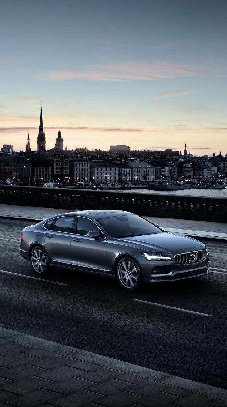 a silver volvo car driving down the road in front of a cityscape at dusk