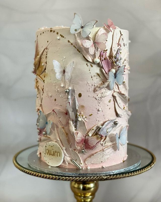 a pink cake with butterflies on it sitting on top of a glass plate next to a gold pedestal