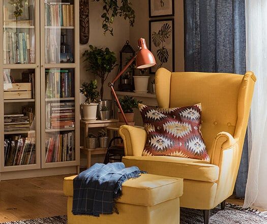 a yellow chair and ottoman in front of a bookshelf with plants on it