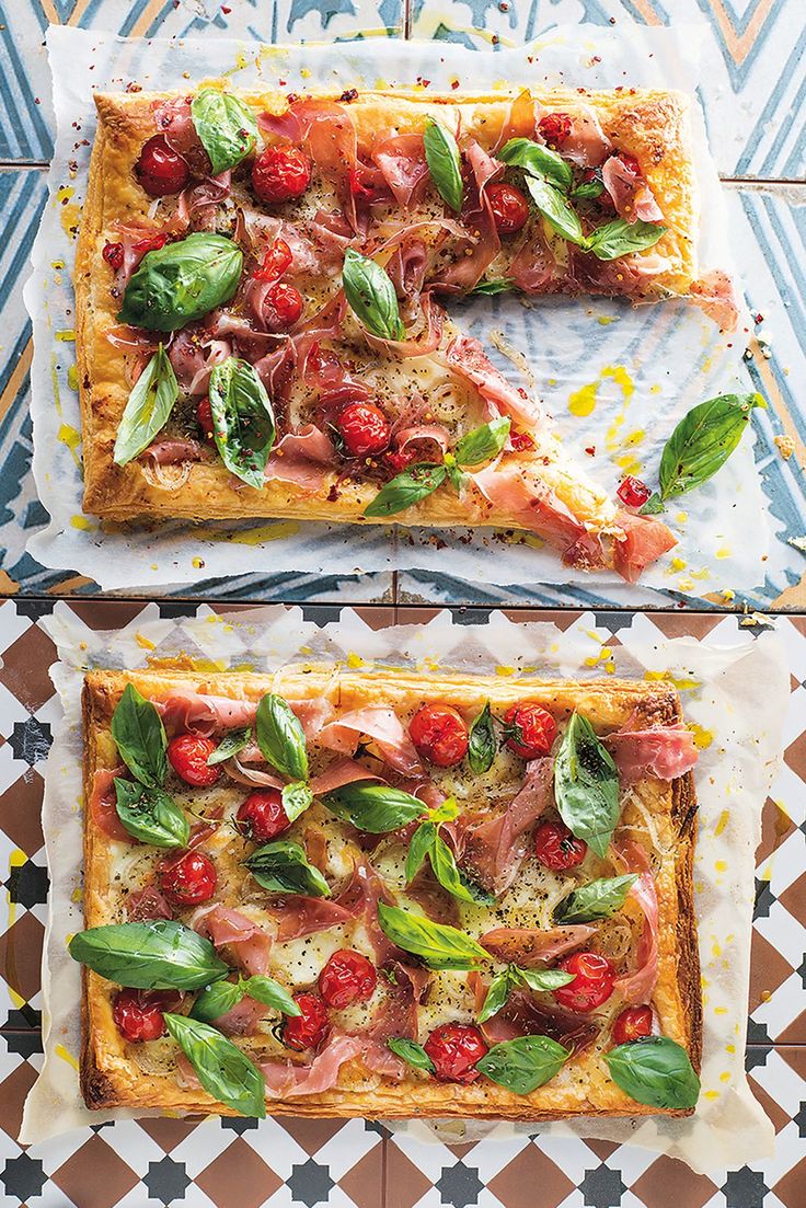 two rectangular pizzas with tomatoes, ham and basil on them sitting on top of a patterned tablecloth