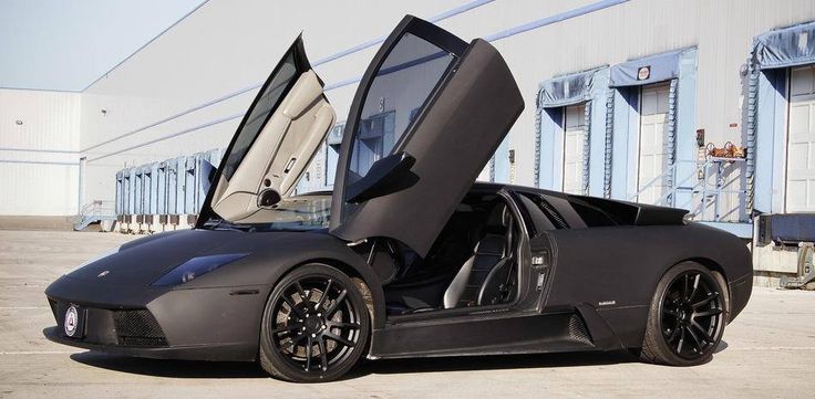 a grey sports car with its doors open in front of a building that has blue shutters