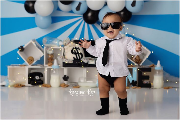 a baby boy wearing sunglasses and a tie standing in front of a backdrop with balloons