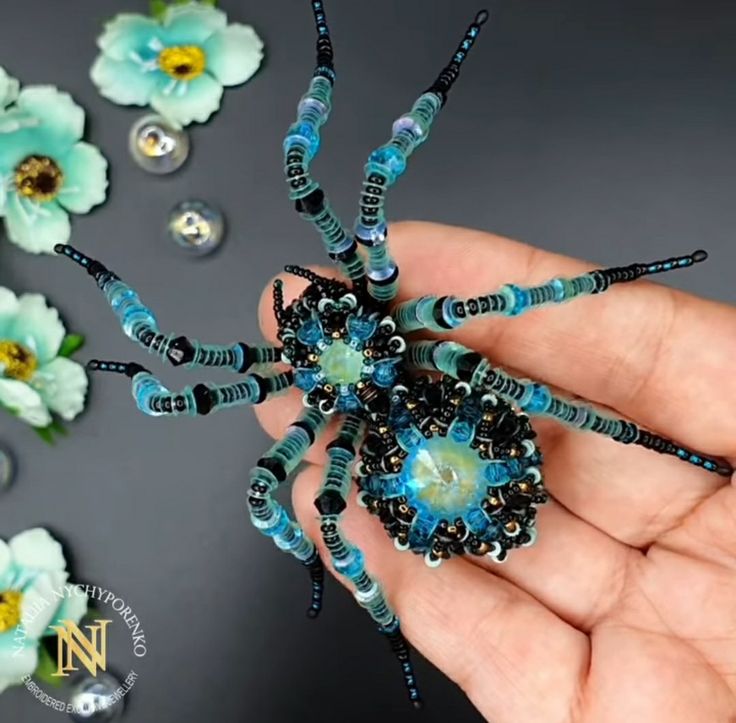 a hand holding a beaded spider with blue beads on it's body and flowers in the background